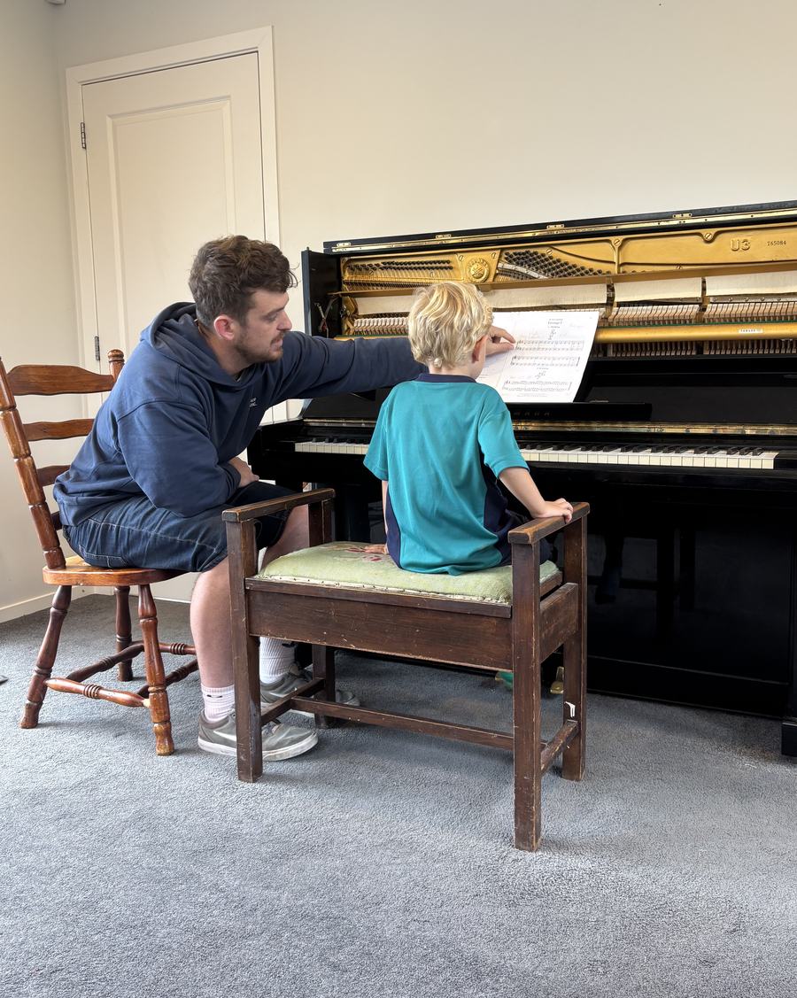 Adam Whybro sitting beside a young student at the upright piano, pointing at the open sheet music while the student plays. The student is seen from behind.