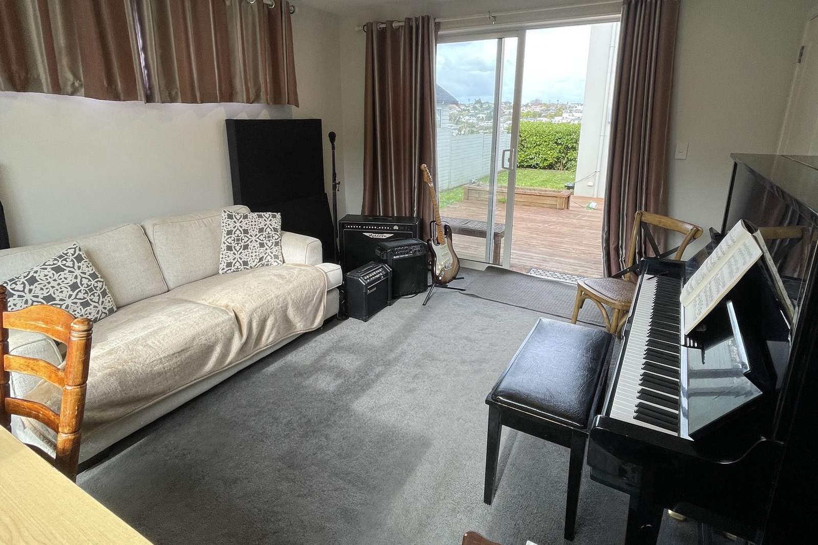 The teaching room at the Long Bay studio, with a sofa on the left, an electric guitar and two amplifiers by the sliding doors, and an upright piano with sheet music open on the right